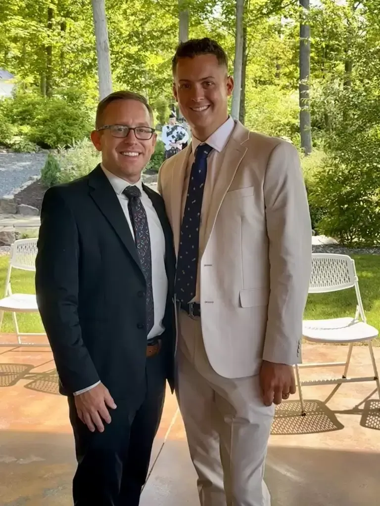 Two men in suits smiling outside. One wears a dark suit, the other a light-colored suit.