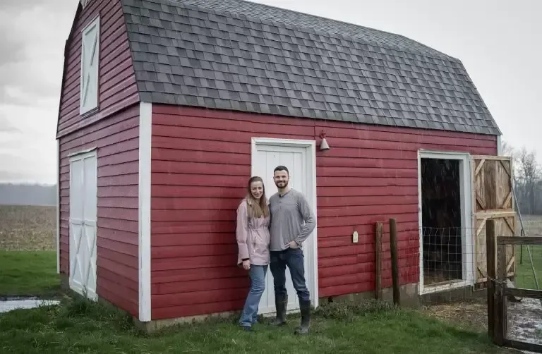 Couple standing in front of a red barn with white trim and a grey roof; they are in a field.
