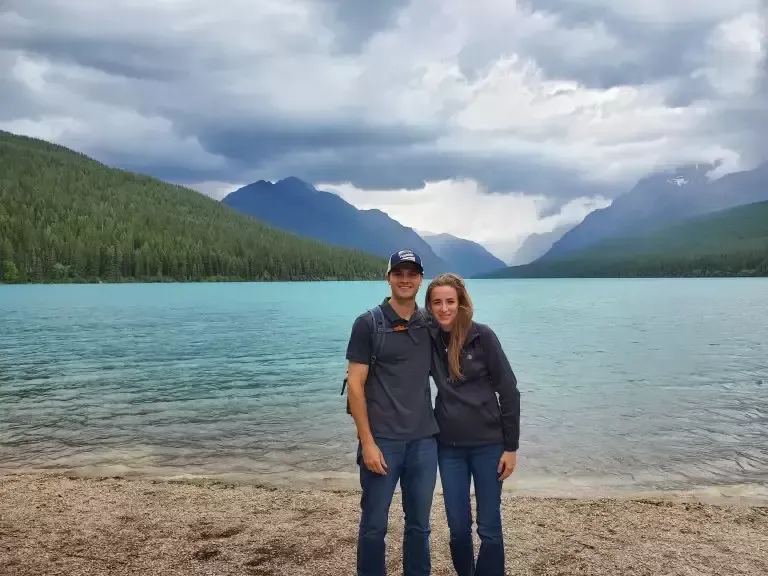 Couple poses by turquoise lake against mountain backdrop, cloudy sky.