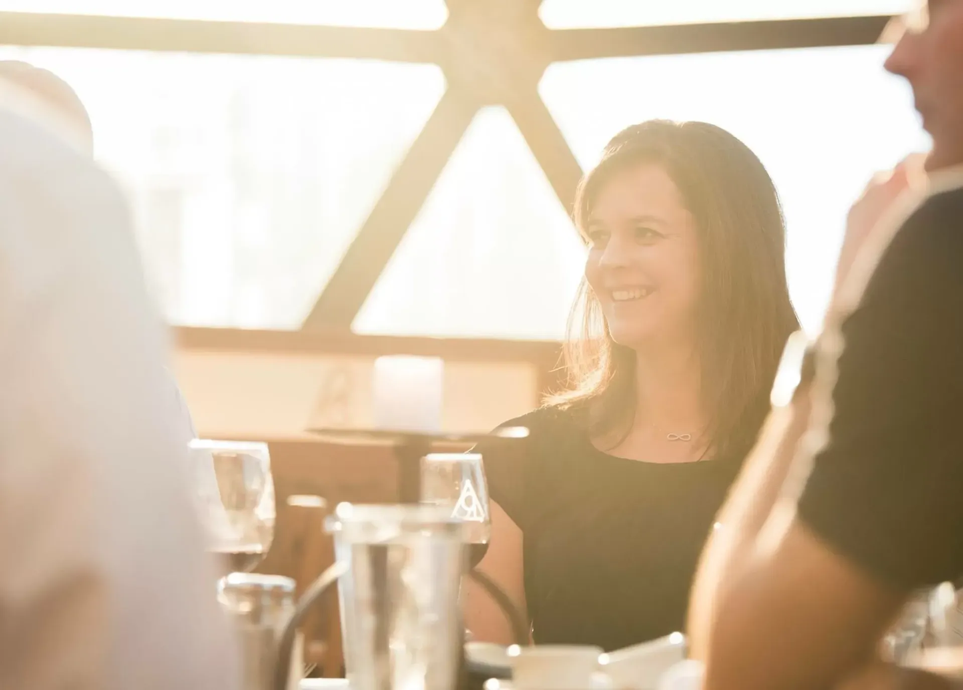 Woman smiling at a table with others; bright light from a window.