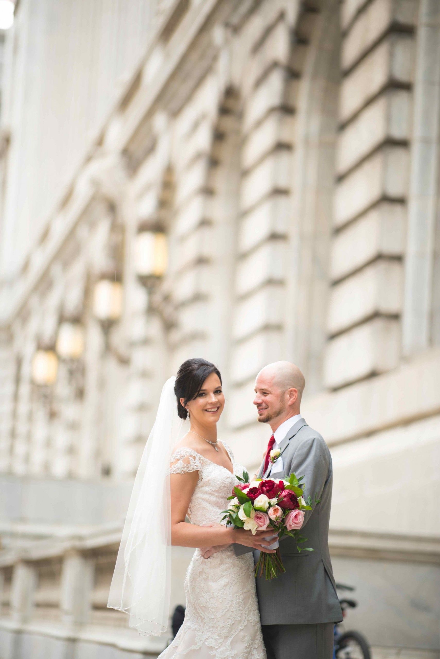Bride and groom smile, holding bouquet, in front of ornate stone building.