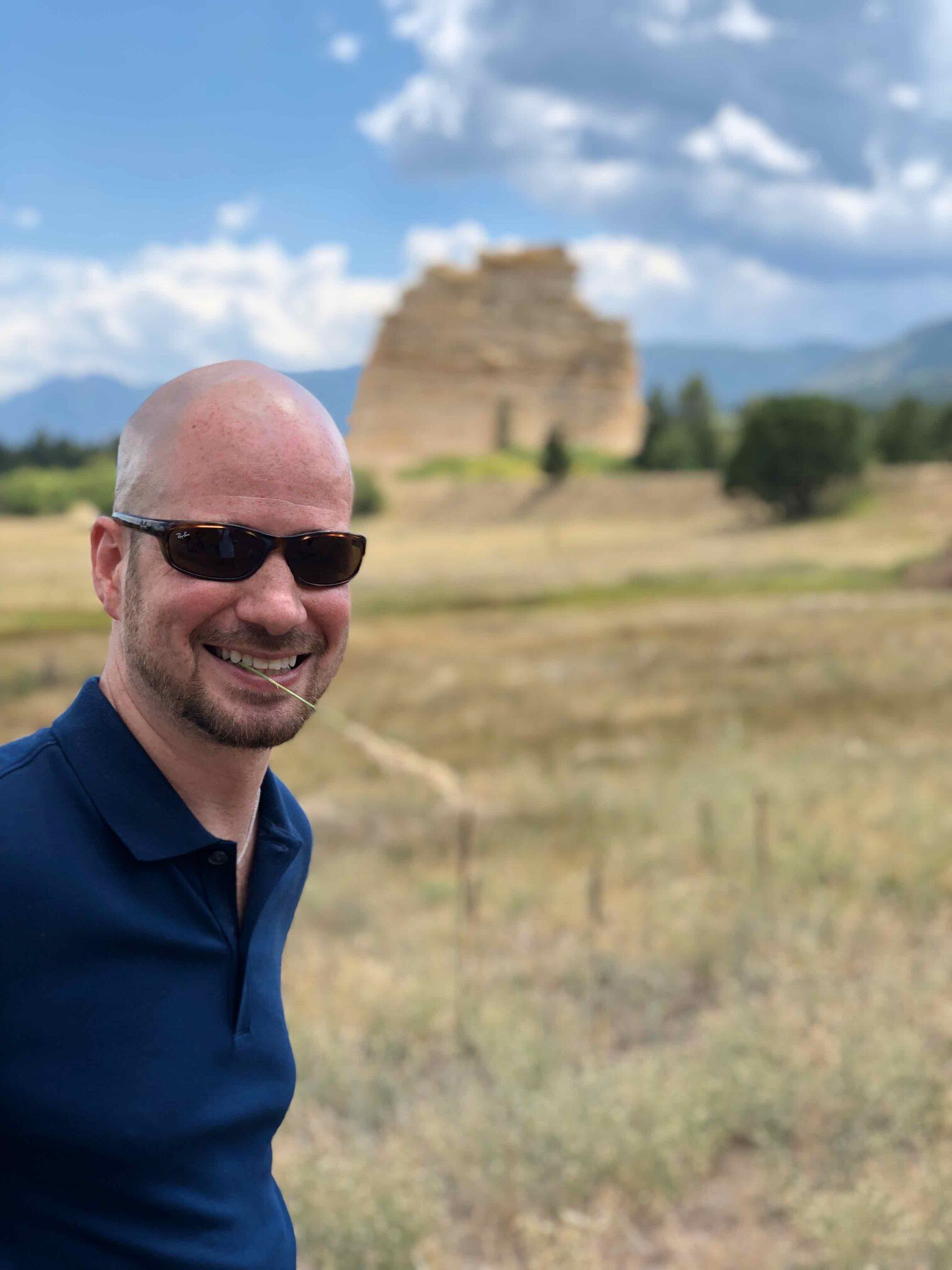 Man in sunglasses smiles in front of a rock formation and field. Blue shirt and sky.