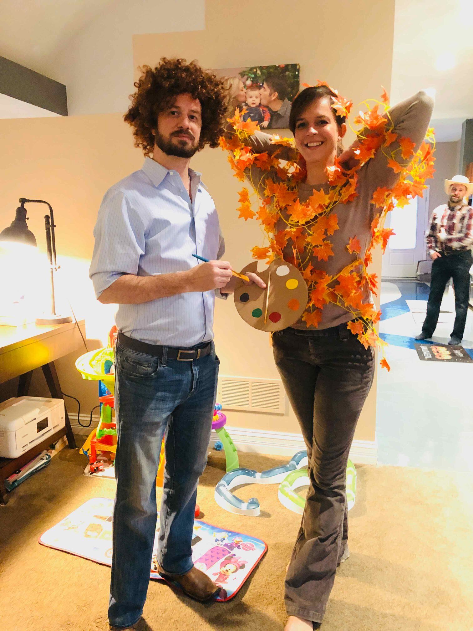 Man dressed as Bob Ross, woman as fall tree, with palette. Inside, bright colors.