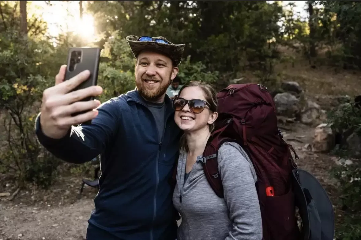 Couple taking selfie on hiking trail; man holds phone, woman smiles with backpack.
