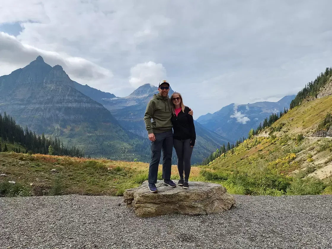 Couple posing on a rock, with mountains and a cloudy sky in the background.
