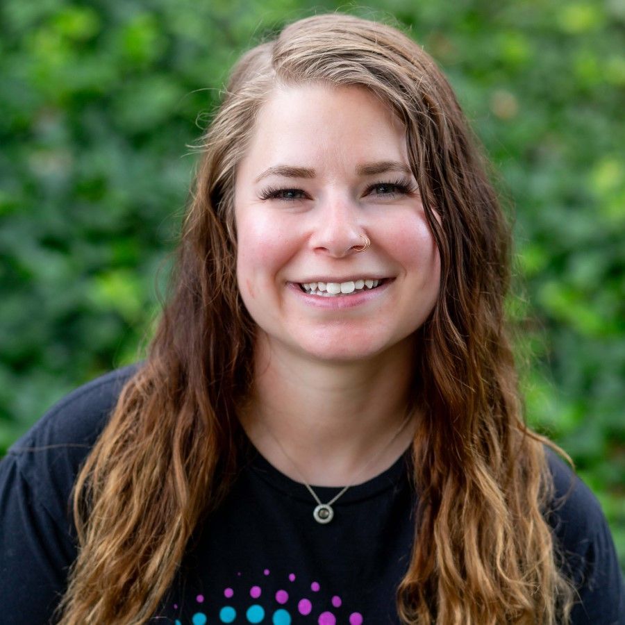 Woman with dark hair smiles, wearing a pink jacket and floral print shirt, with a logo that says 