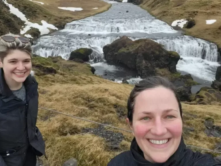 Two smiling women pose near a multi-tiered waterfall in Iceland, with snow patches and brown vegetation visible.