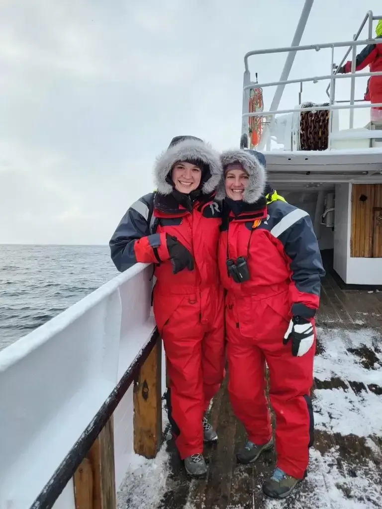 Two people in red winter suits pose on a snowy ship deck, ocean in the background.