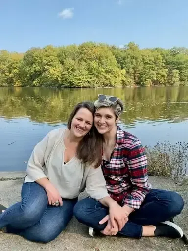 Two women smiling, sitting by water, in front of a tree-lined shore.