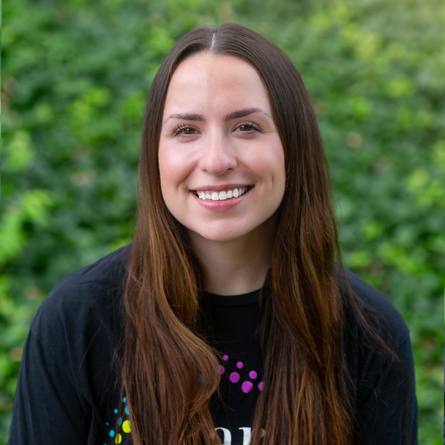 Woman with long brown hair smiles at the camera, wearing a black shirt with colorful dots. Green background.