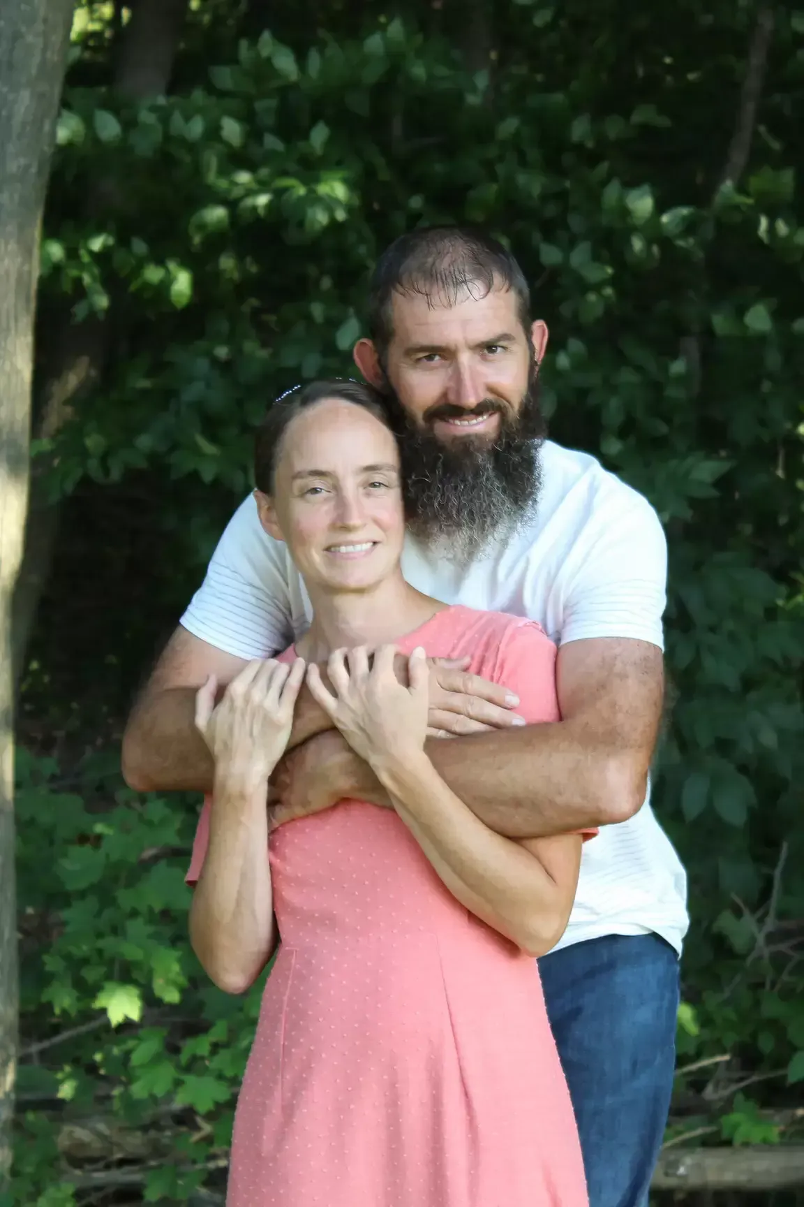 Couple embracing outdoors; woman in pink dress, man with beard in white shirt.