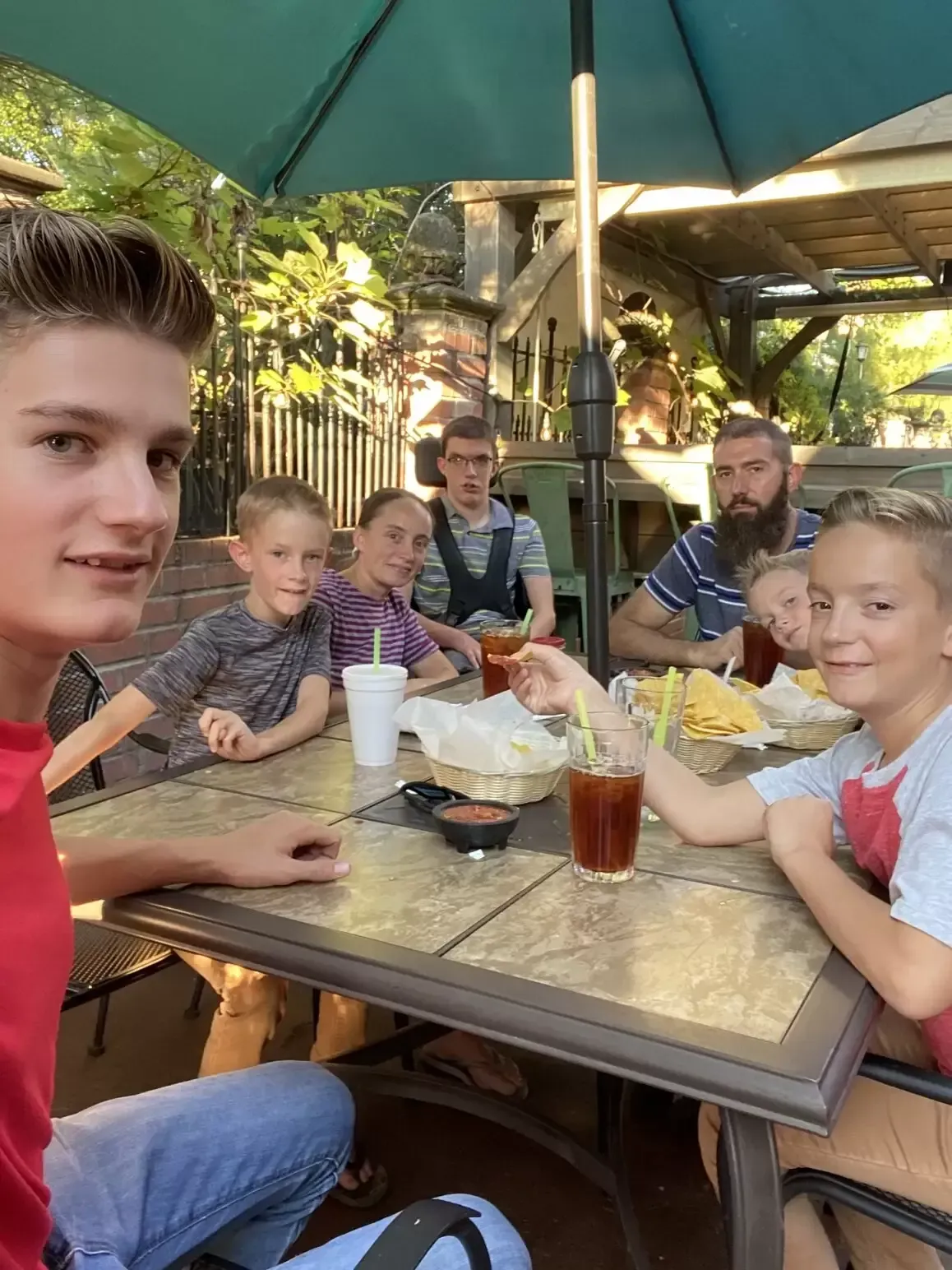 Family seated at outdoor restaurant table, eating chips and salsa. People smiling, under an umbrella.