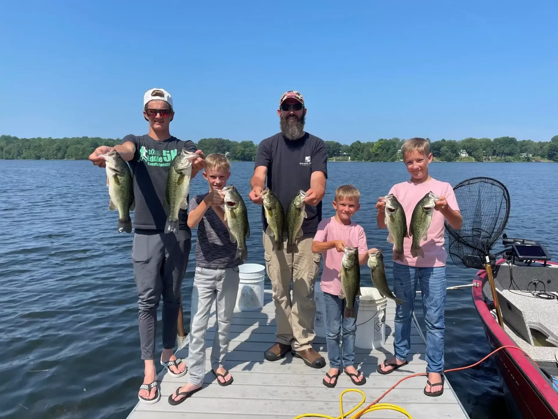 Five people holding up fish on a dock. Sunny day, lake in background.