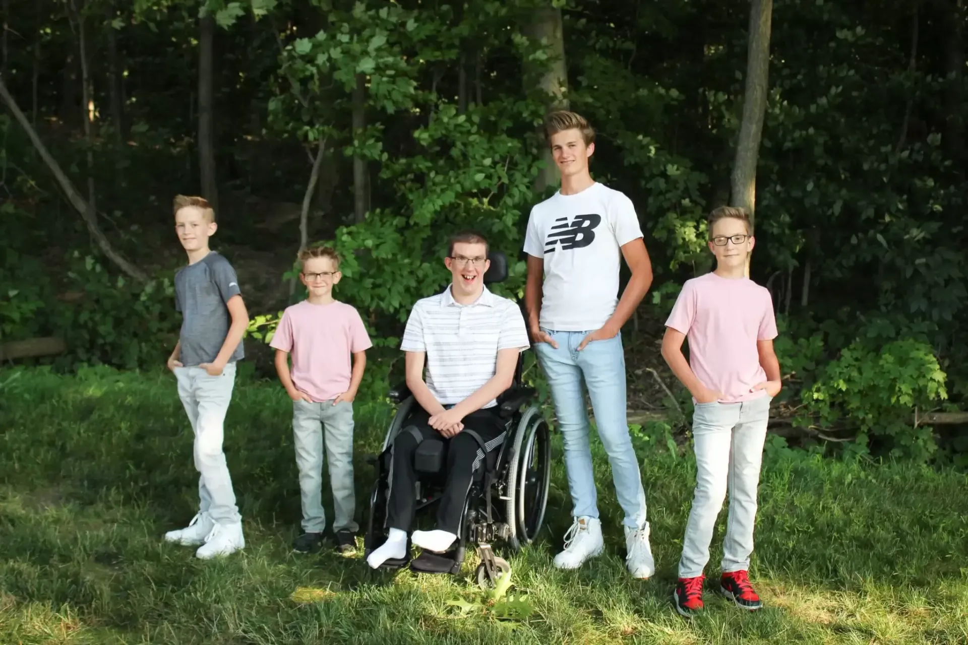 Five boys pose outdoors; one in a wheelchair. Green trees in background.