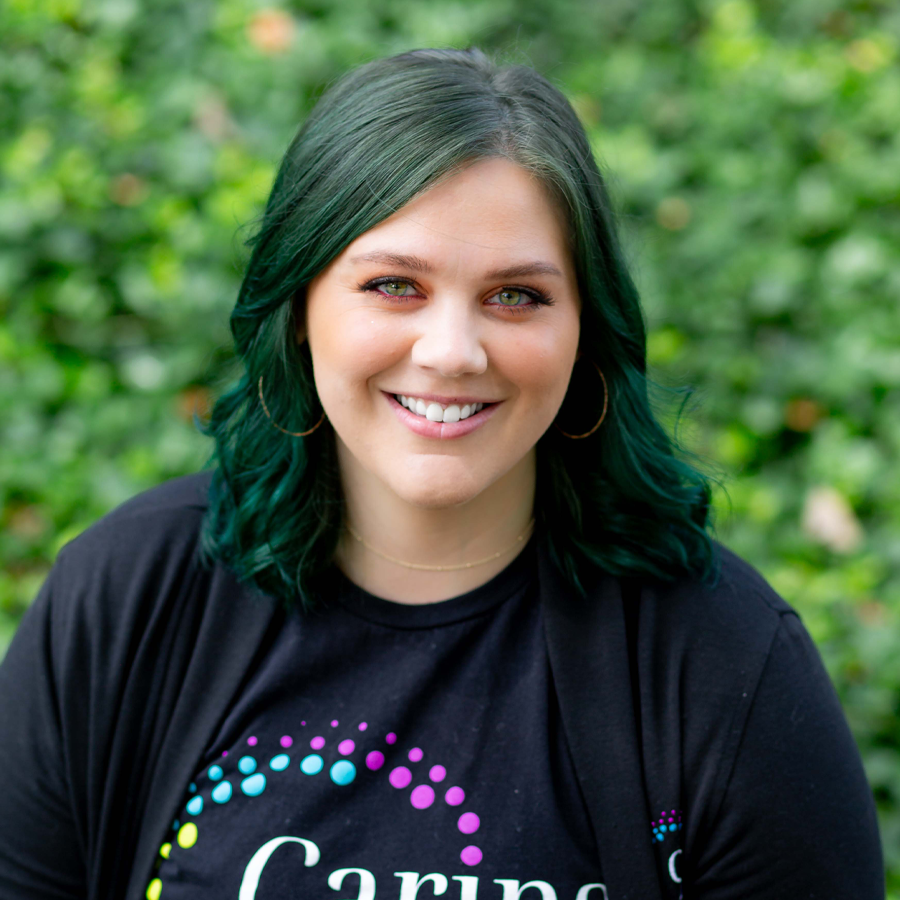 Woman with green hair smiles, wearing a black shirt in front of green foliage.