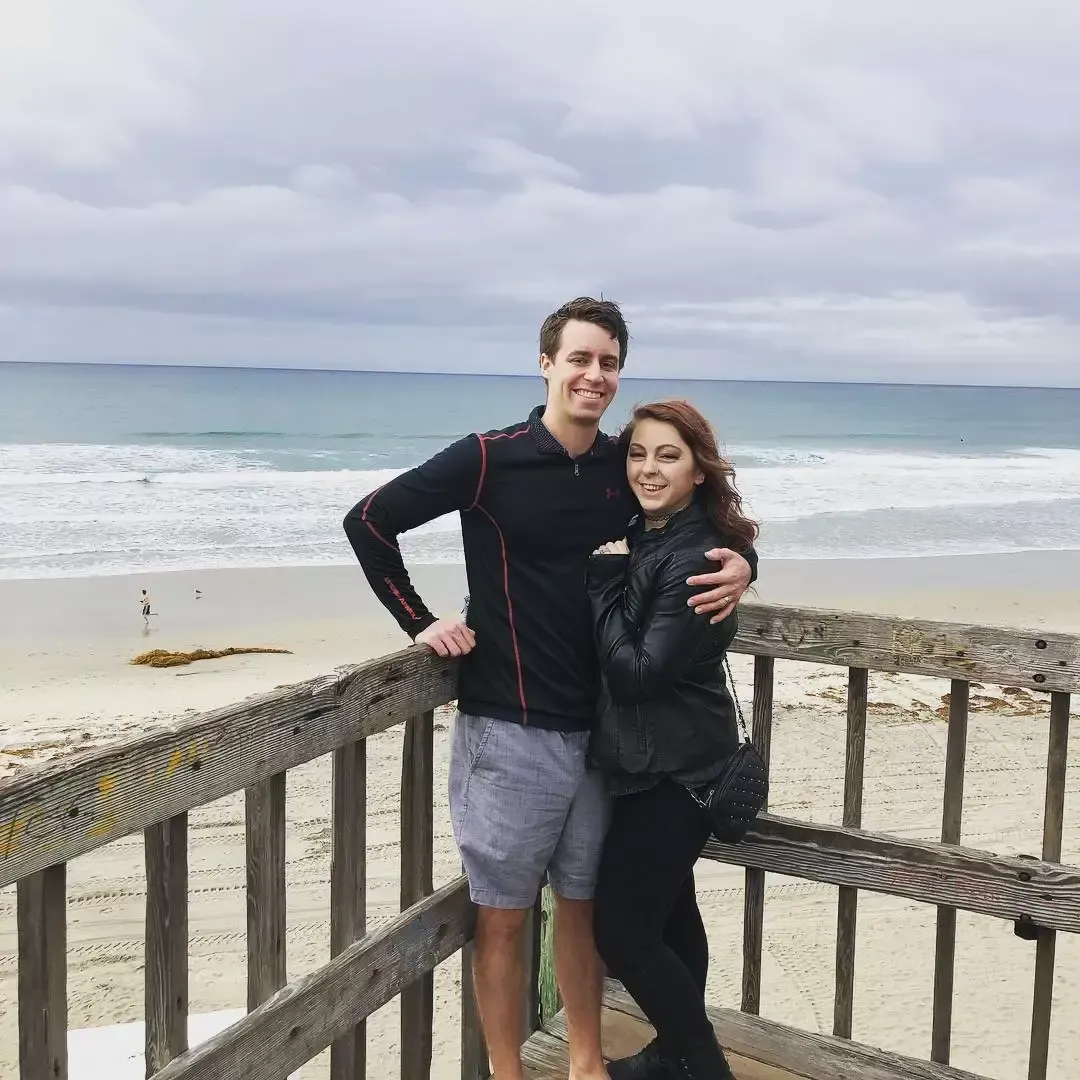A couple stands on a wooden deck by the beach, smiling. Man has arm around woman. Cloudy day.