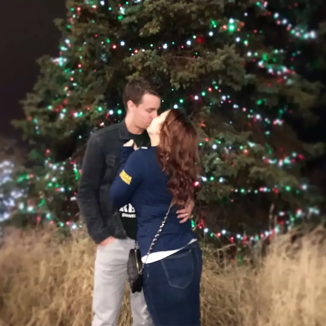 Couple kissing in front of a Christmas tree with colorful lights at night.