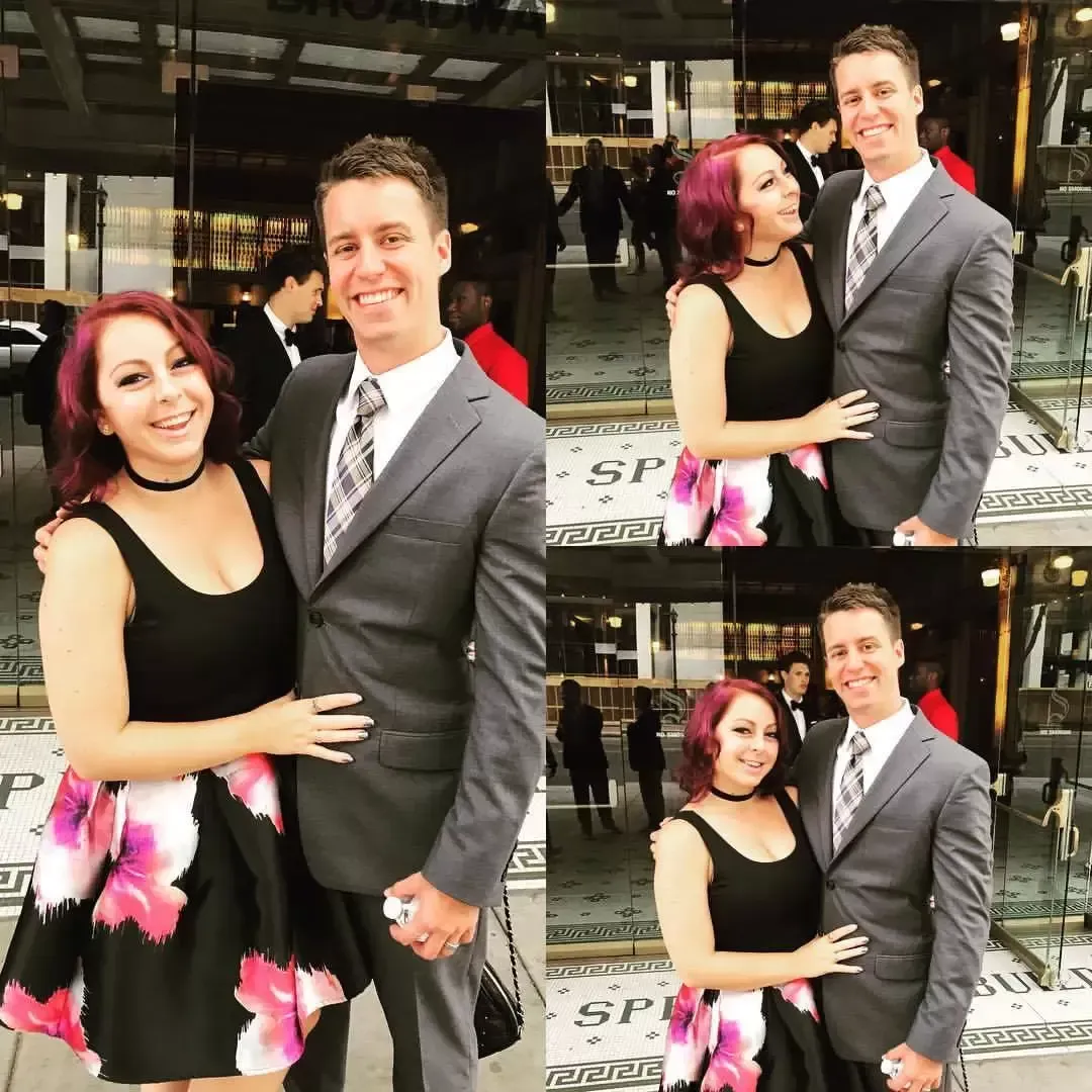 A man and woman pose outside. The woman wears a black dress with pink flowers, and the man wears a gray suit.