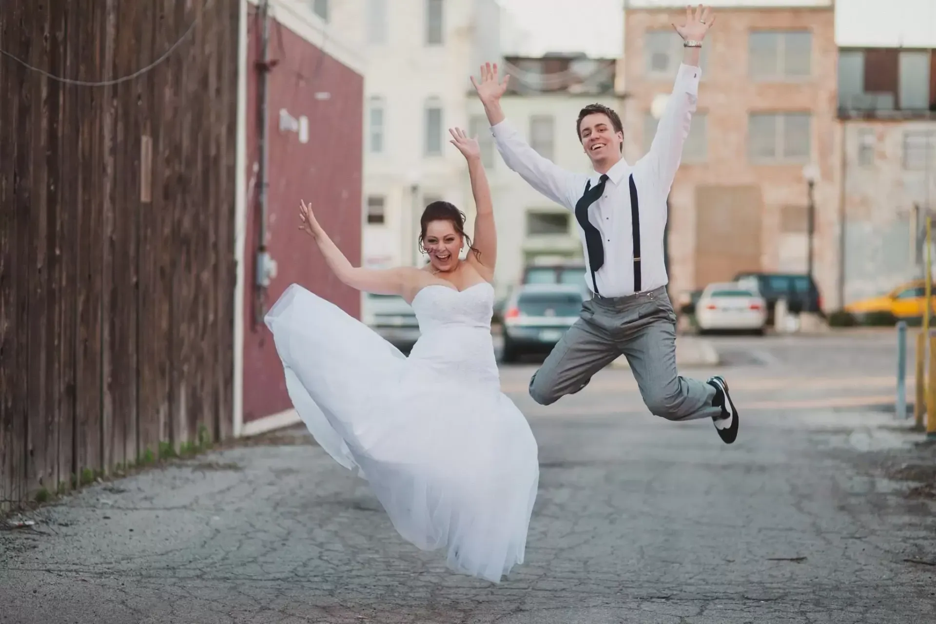 Newlyweds jump for joy in alley, woman in white dress, man in suspenders, arms raised.