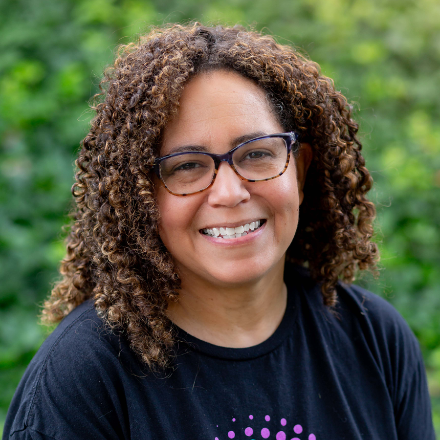 Woman with curly brown hair, glasses, smiling in front of a green background.