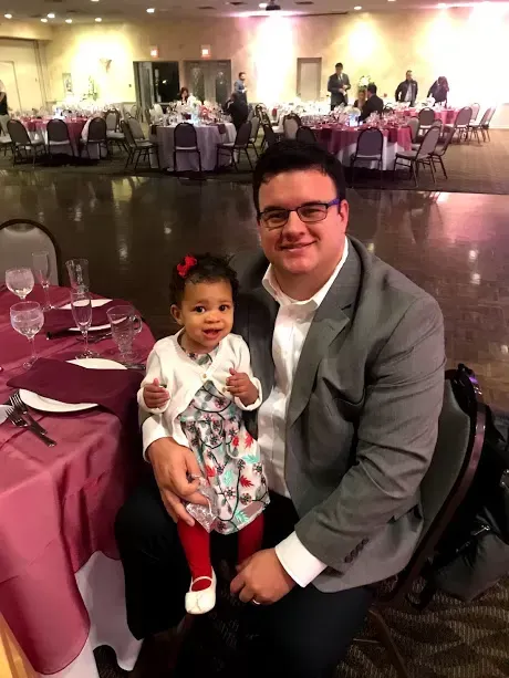 Man in glasses with a young girl in a dress at a formal event, sitting near a table.