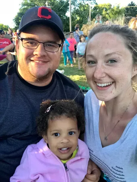Family selfie: smiling man in cap, woman, and young child in pink jacket outdoors on grass.