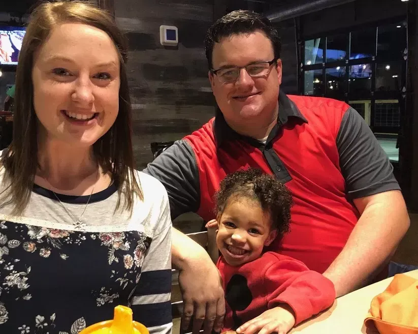 A woman, man, and child smiling at the camera. They are indoors and the child is in front of the man.