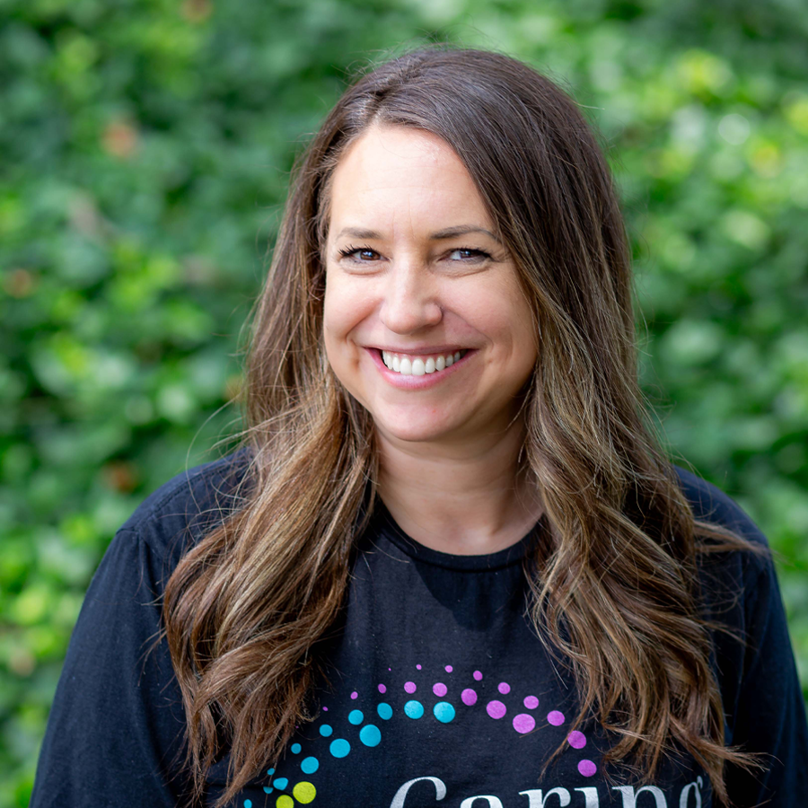 Woman with long brown hair smiling, wearing a black shirt with colorful dots, set outdoors.