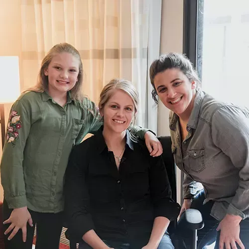 Three women smiling in a room. One sits in a chair, two stand, one with arm around her.