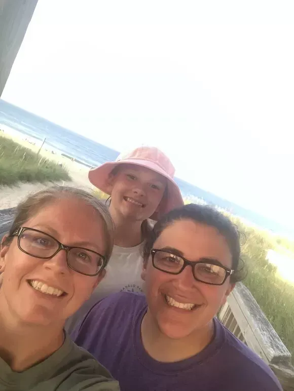 Three smiling people near a beach: two adults wearing glasses, one child in a pink hat.