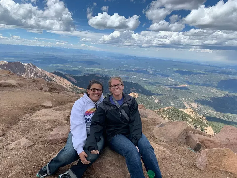Two women sitting on a rocky mountaintop, smiling, with a vast valley and cloudy sky in the background.