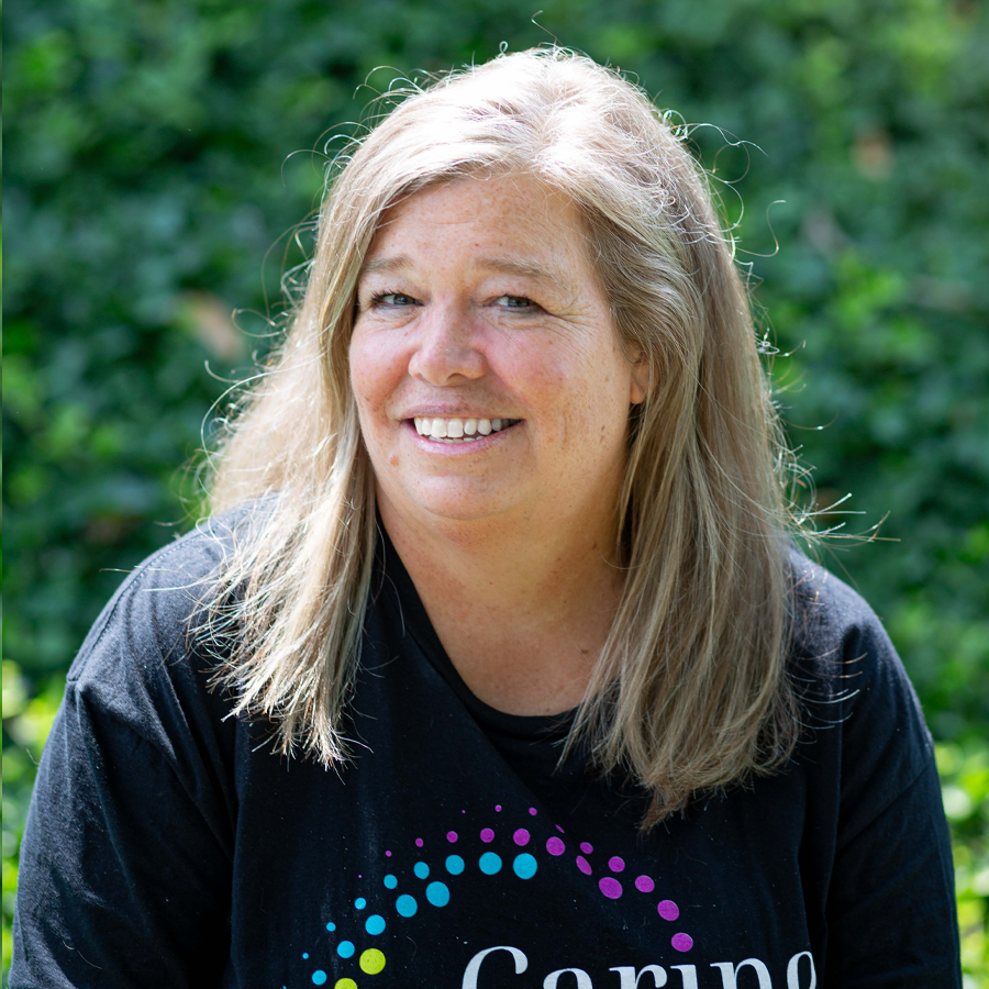 Woman with blonde hair smiles, wearing a black shirt with a colorful design, set outdoors in front of green foliage.