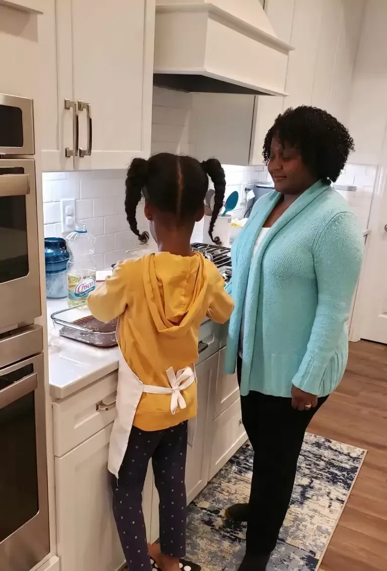 Girl in apron baking with an adult in a kitchen, white cabinets, blond countertop.