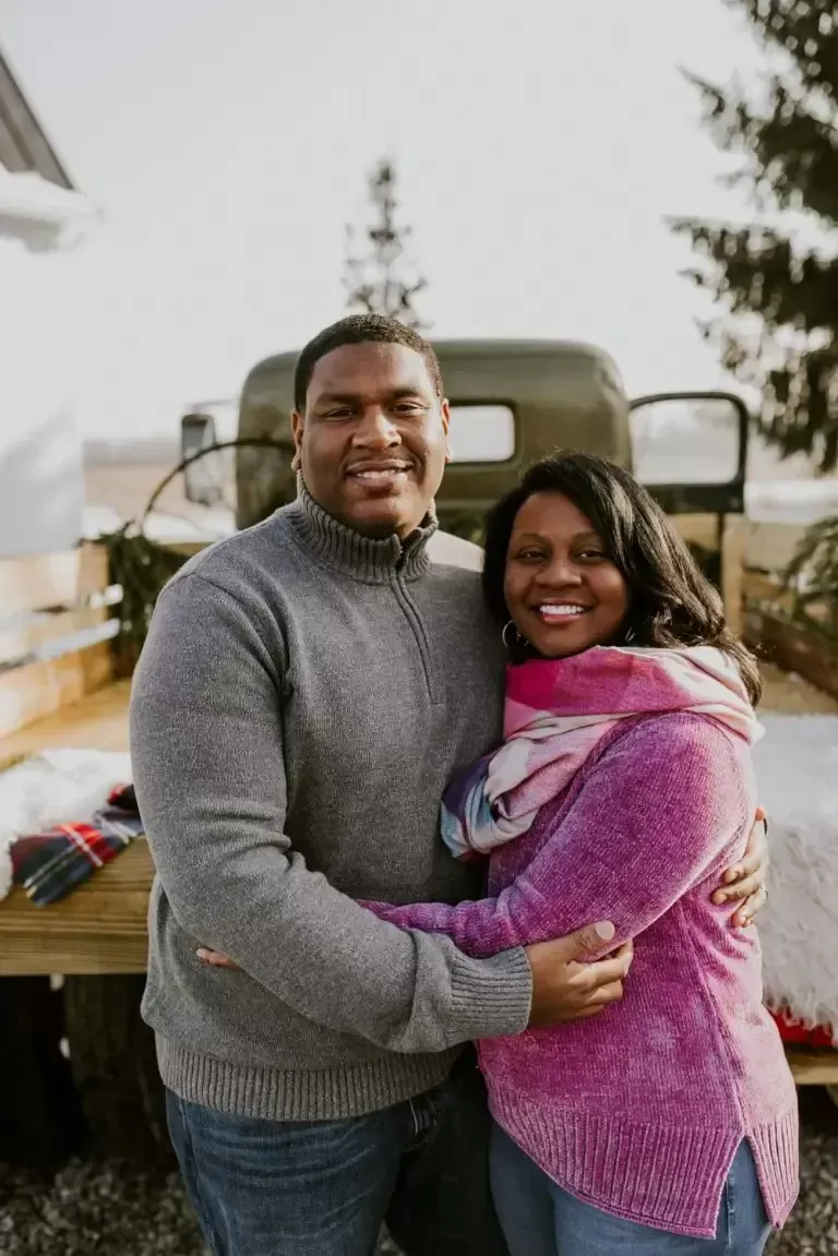 Couple hugging in front of an old truck with a winter scene, both smiling.