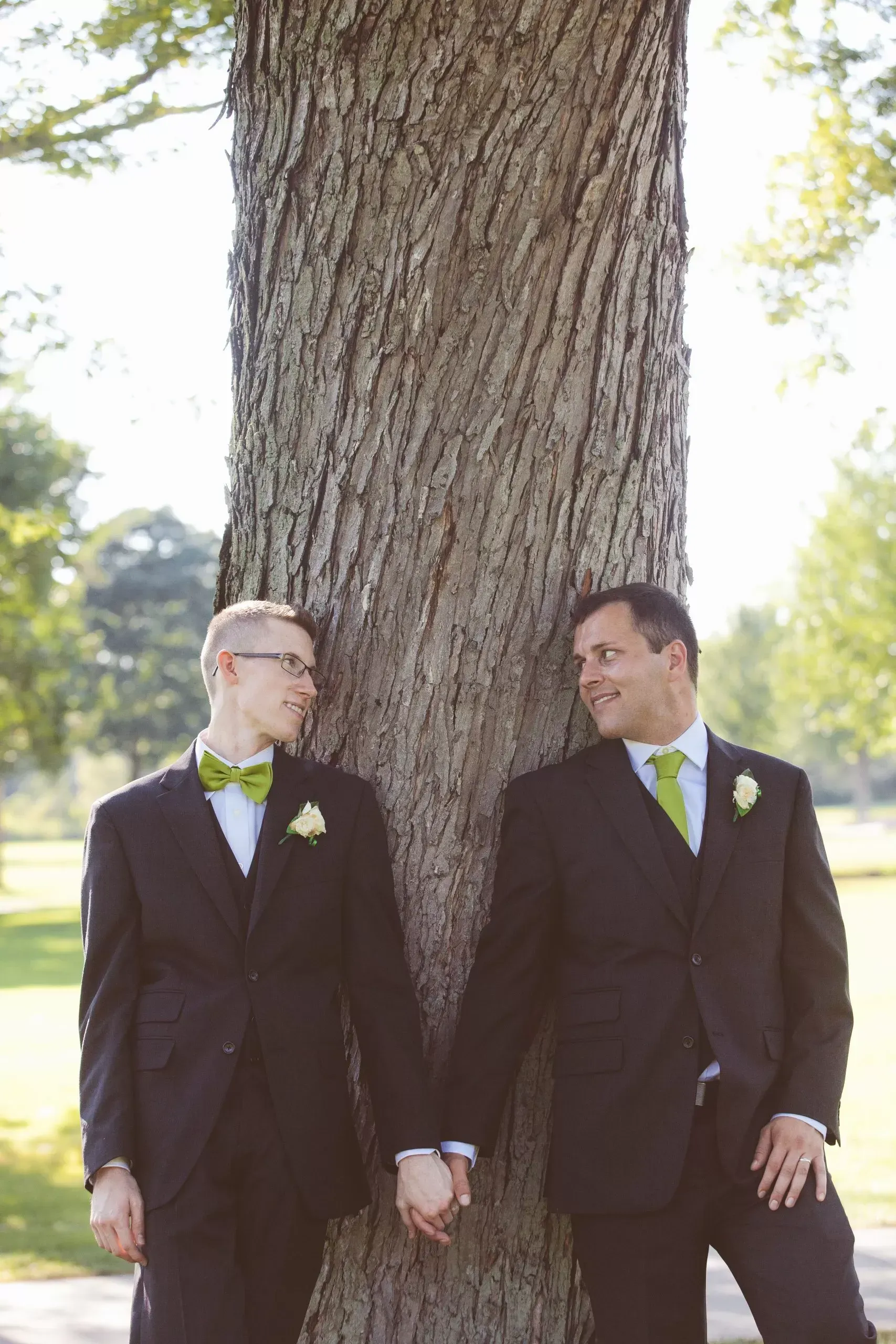 Two men in suits holding hands, leaning against a tree, smiling at each other, outdoors.