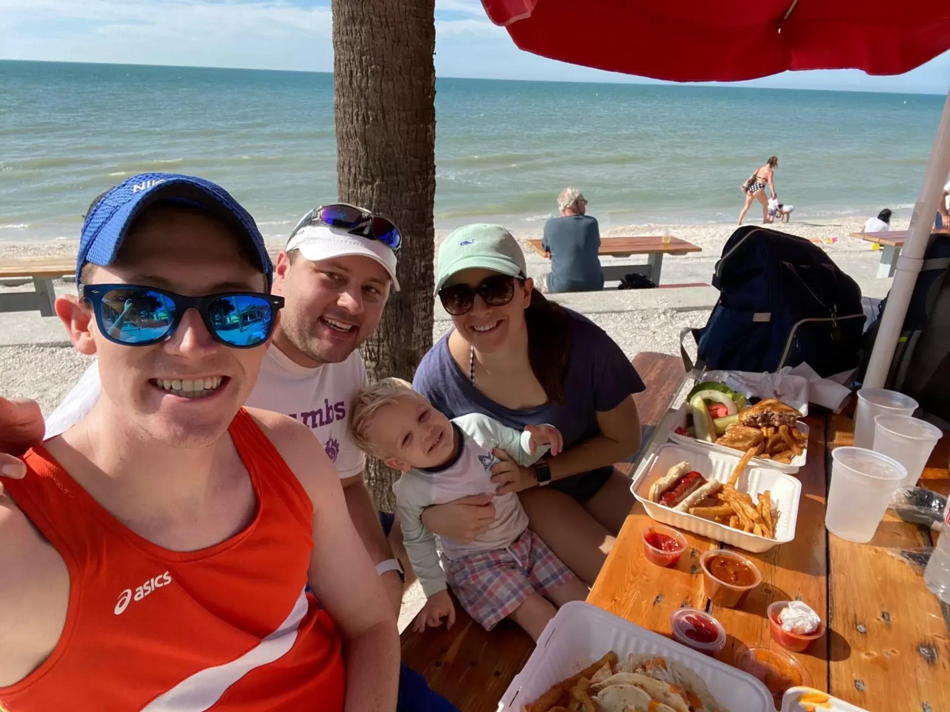 Group of four smiling people and a child at a beach table with food, under shade; ocean in background.