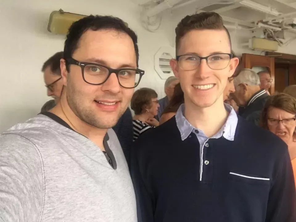 Two men smiling, wearing glasses, on a cruise ship deck with other passengers in the background.