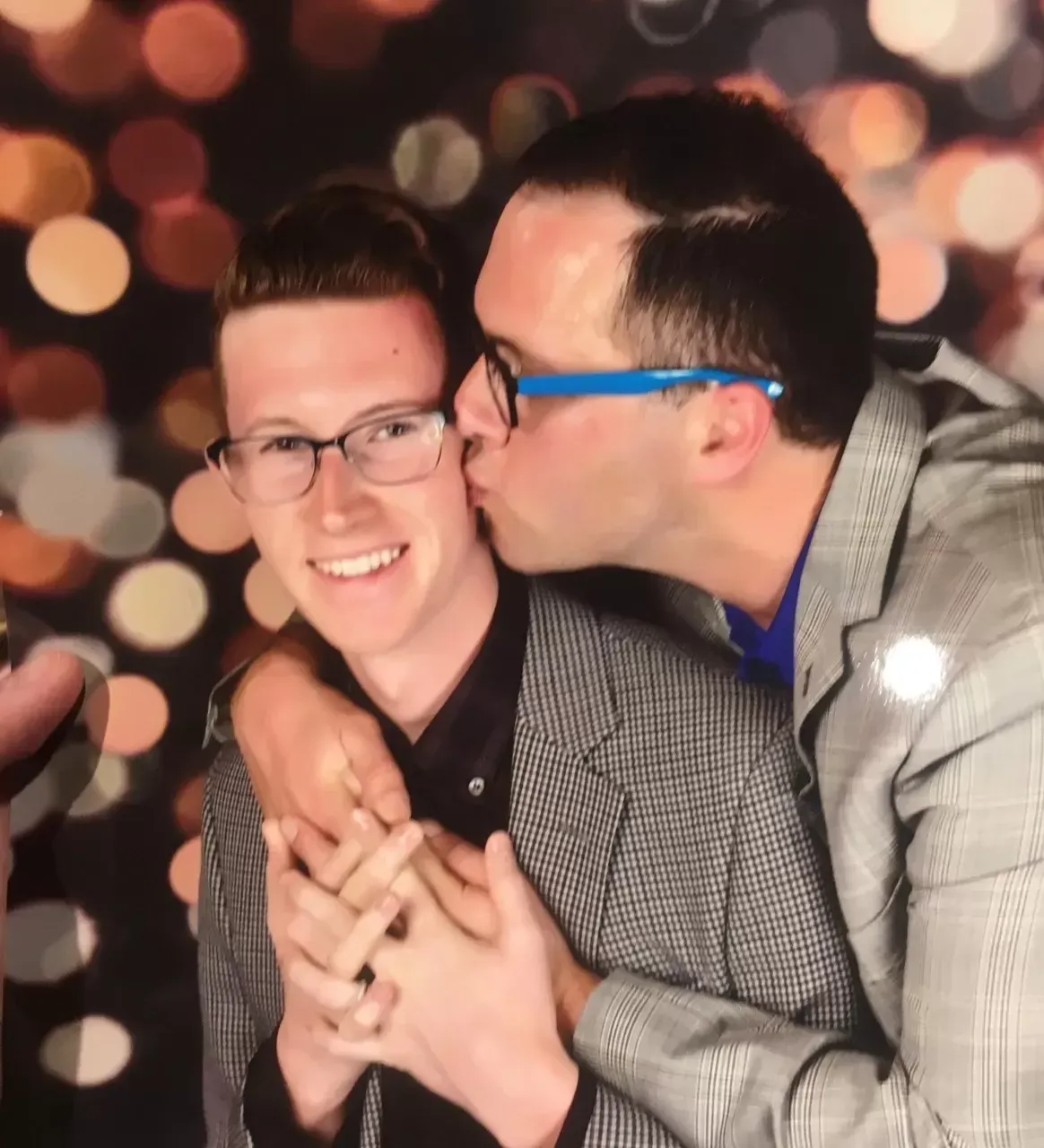 Two men in suits, one kissing the other's cheek. Both wear glasses, smiling against a bokeh background.