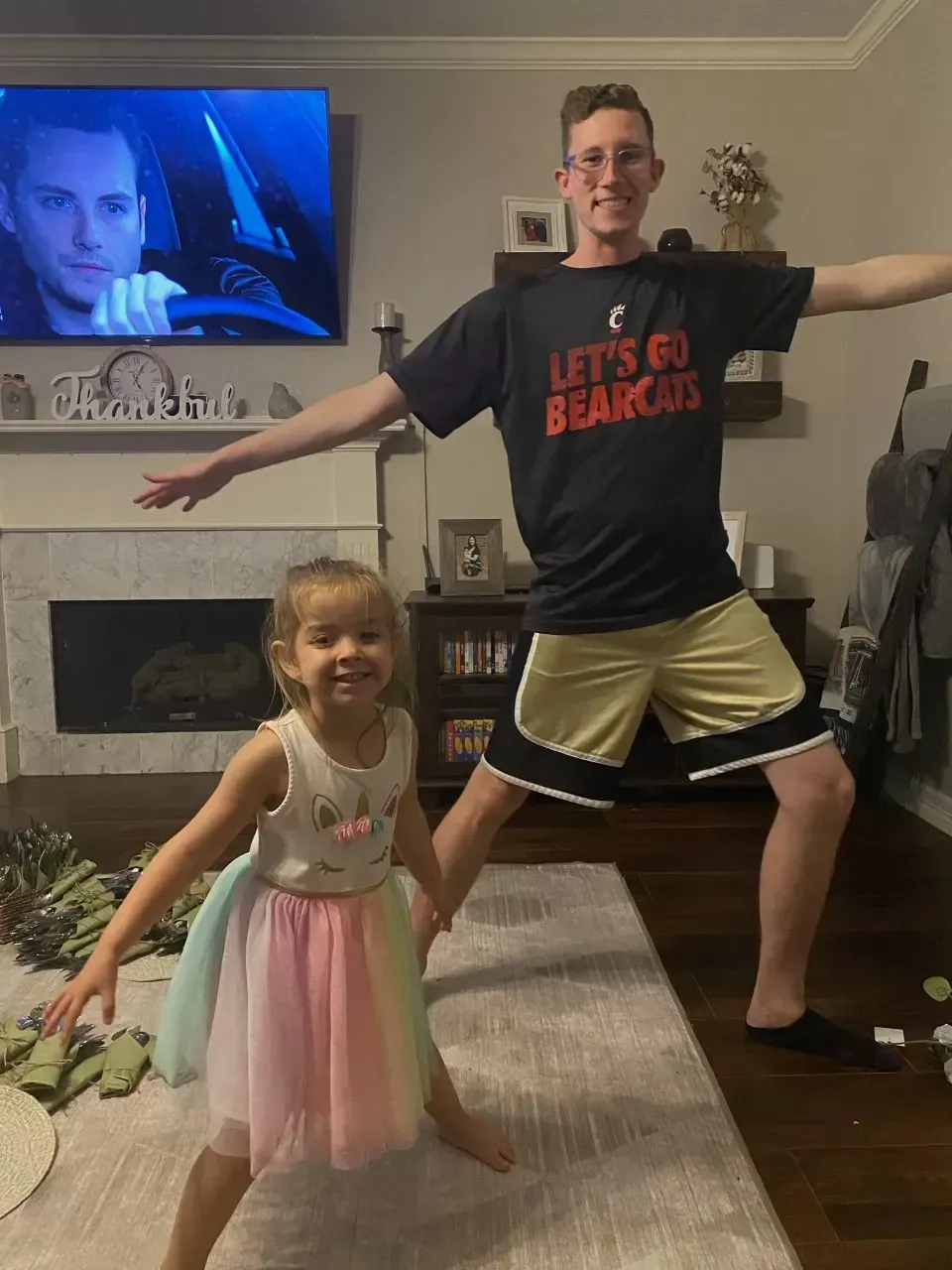 Man and young girl in home pose in athletic stance; girl wears colorful tutu, man wears Bearcats shirt.