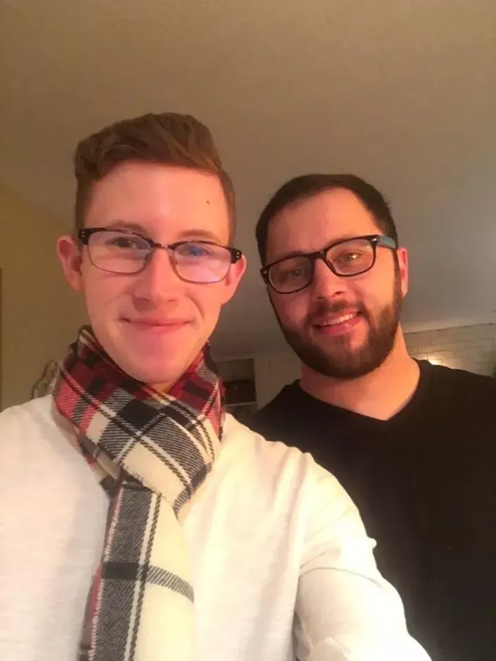 Two men smiling for a selfie; one wears glasses and a plaid scarf, the other a black shirt.