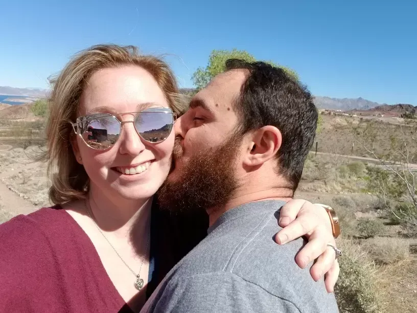 Smiling woman with mirrored sunglasses, being kissed on the cheek by a bearded man, outdoors in a desert landscape.