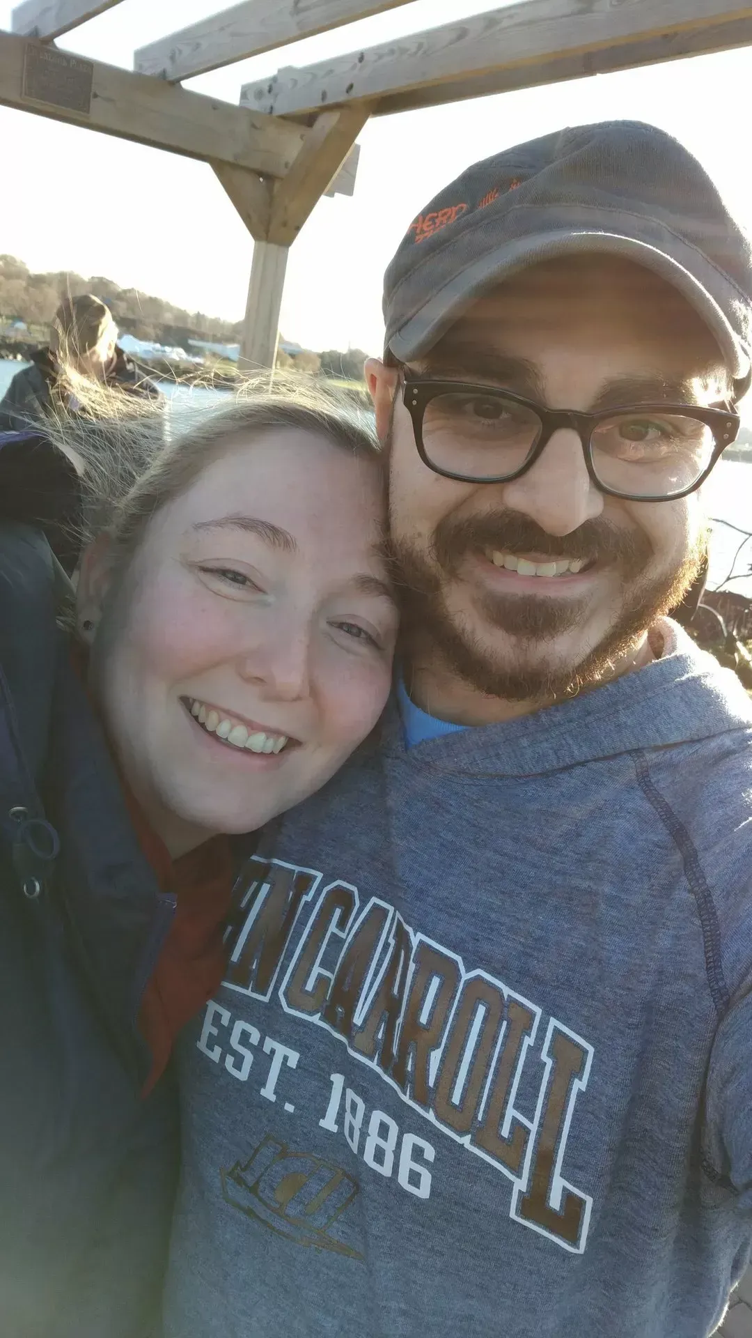Woman and man smiling, close-up selfie. Man wears glasses and a cap, woman's head on his shoulder. Outdoors, sunny.