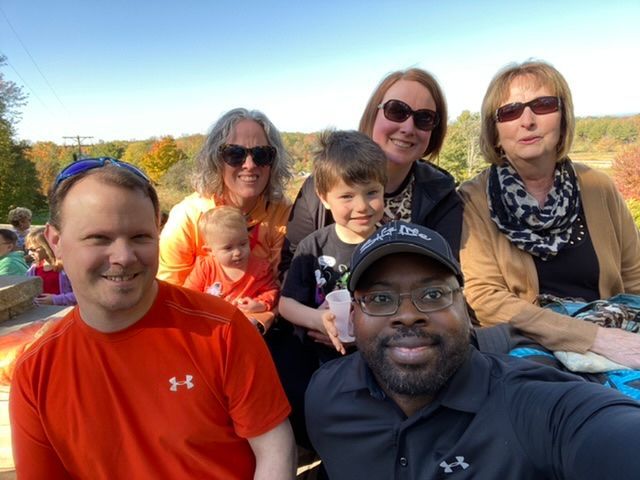 Group selfie outdoors; people smiling; autumn foliage in background.