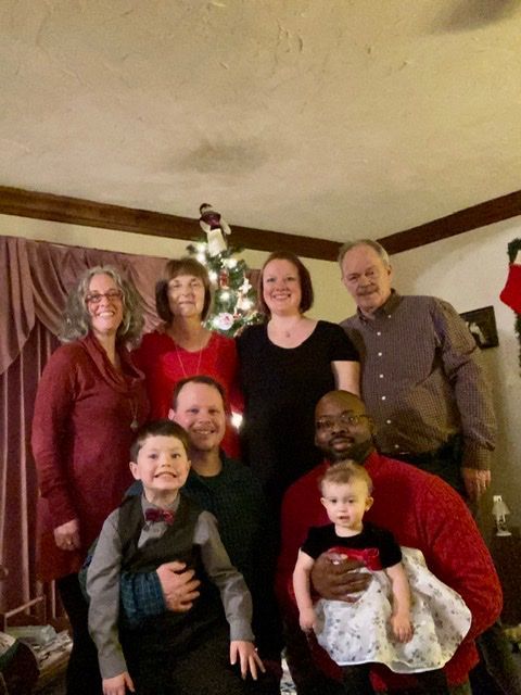 Family portrait in front of a Christmas tree. Adults and children smiling, wearing festive colors.