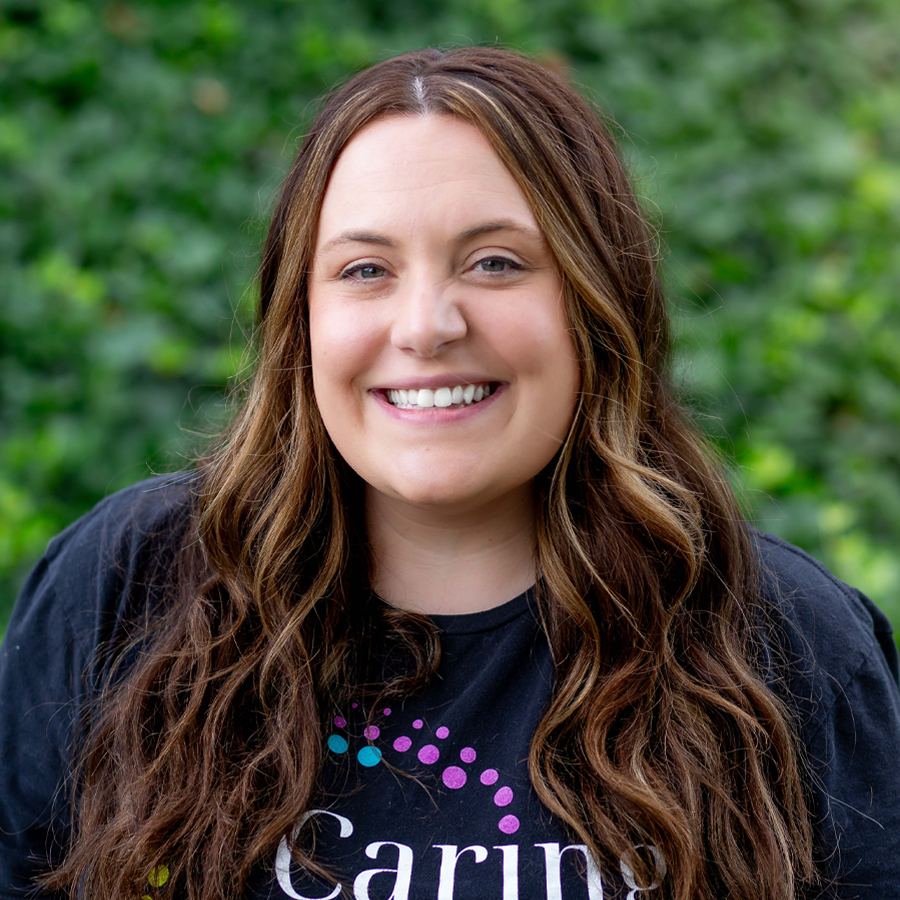 Woman with brown hair smiles, wearing a black shirt in front of a green leafy background.