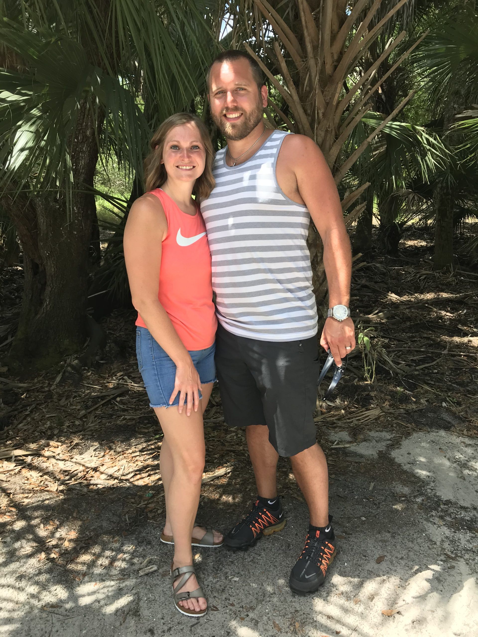 Woman in orange shirt and denim shorts stands with man in striped tank top and shorts outdoors, smiling.