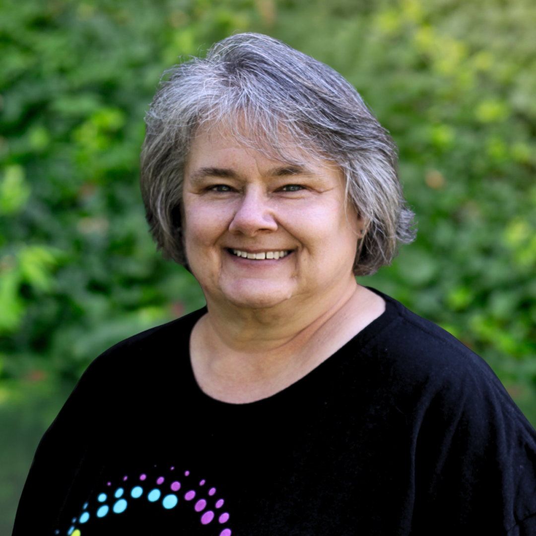 Woman with glasses smiles, wearing a floral shirt against a gray background.