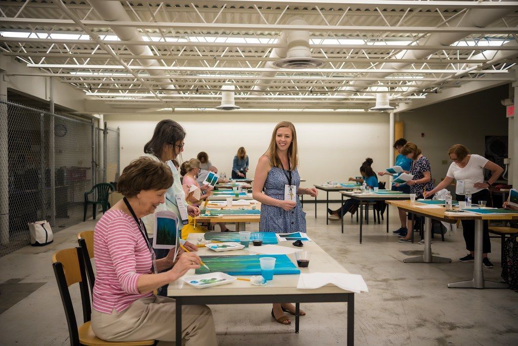 People craft at tables in a brightly lit room. A woman in a pink shirt works closely, and another assists.