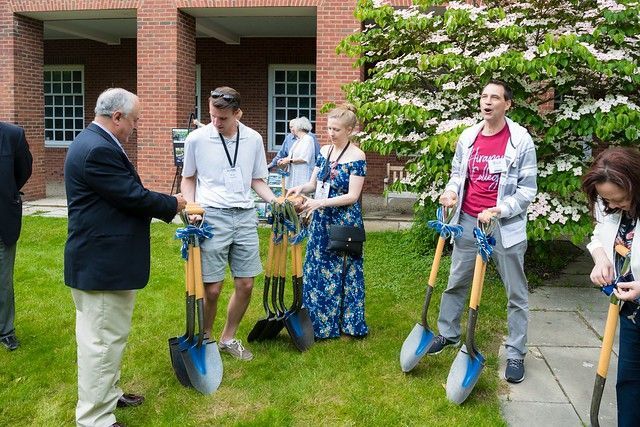 People with shovels gathered on grass, preparing for a ground-breaking ceremony near a brick building.