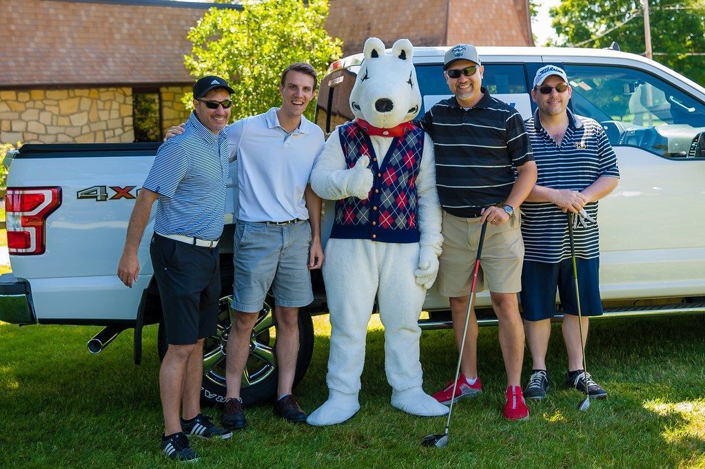 Group of men and a polar bear mascot pose with a truck and golf clubs on a grassy lawn.
